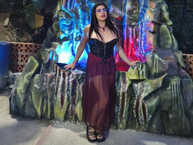 KhalifaGold se encuentra frente a una escultura rocosa, iluminada con colores vibrantes y luces de neón. Ella lleva una blusa negra de encaje y una falda burdeos transparente, mostrando el escote y un collar. Es atractiva, con un aspecto increíblemente sexy..