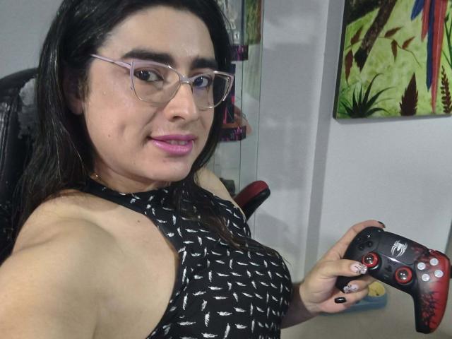 KhalifaGold with black hair and glasses, wearing a black patterned sleeveless top, holding a red and black gaming controller, looking at the camera with a slight smirk. Casual, intimate selfie.