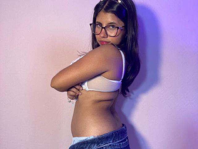 Fotografia di CelesteMorel, una donna formosa con lunghi capelli castani, che indossa occhiali e un reggiseno bianco, e che guarda verso l'alto. la spalla scoperta, la mano sul petto, jeans blu leggermente abbassati.