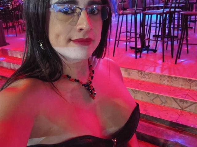 KhalifaGold, vestida con un top negro sin tirantes, gafas y un collar de cuentas negras, está sentada en un bar iluminado con luces de neón, con colores rojo y luces azules.
ella tiene el pelo largo y negro y una sonrisa segura..