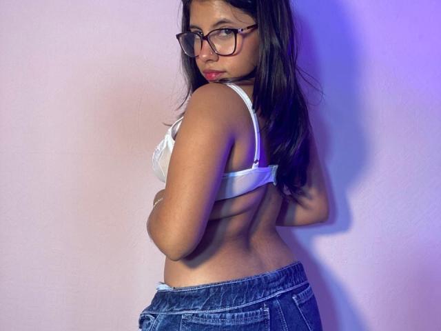 Fotografie von Celeste Morel mit langen, schwarzen Haaren, die eine Brille trägt, ein weißes BH-Oberteil und blaue Jeans anhat und über die Schulter schaut. Hand auf dem Rücken, verführerischer Ausdruck, blau-weißes Licht, sexy Atmosphäre..