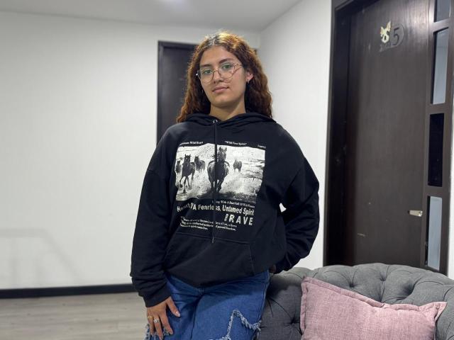 TaniaLux, curly brown hair, glasses, black hoodie with white graphic, blue jeans, standing in modern room, grey couch, pink pillow, dark wood door, casual pose, slight smirk, sexual tension.