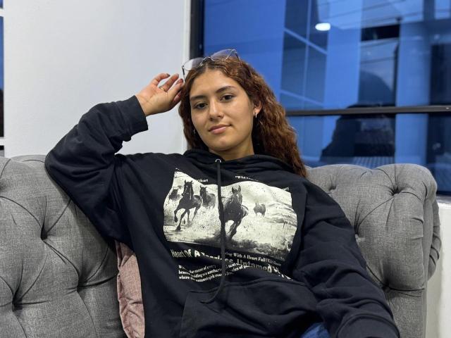 TaniaLux, con i capelli castano ricci, indossa una felpa nera con una grafica bianca e si rilassa su un divano grigio imbottito. Sta appoggiando la mano sulla testa, trasmettendo una calma sicurezza..