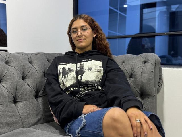 TaniaLux zit op een grijze, gestoffeerde bank, gekleed in een zwarte hoodie met een witte opdruk en gescheurde denim shorts. Ze heeft krullend bruin haar, een bril en een ring aan haar rechterhand. Achtergrond in blauwtinten.