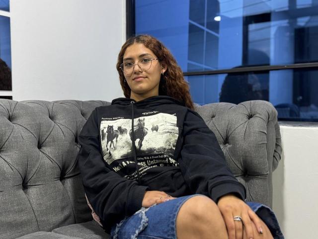 TaniaLux sitzt auf einem grauen, gepolsterten Sofa und trägt eine schwarze Kapuzenjacke mit weißem Aufdruck und Jeansshorts. Sie trägt eine Brille, hat lockiges, braunes Haar und einen Ring an ihrer linken Hand. Eine unglaublich entspannte Atmosphäre..