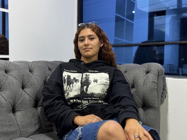 TaniaLux, a woman with curly brown hair, sits on a gray tufted couch, wearing a black hoodie with a white graphic and denim shorts. Blue light fills the background.