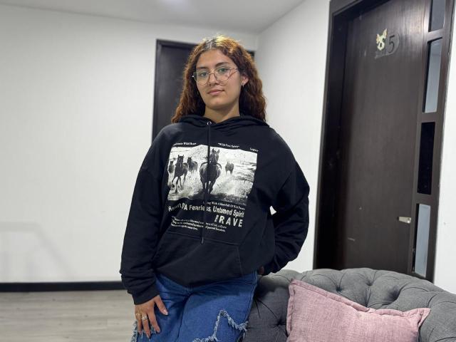 TaniaLux staat in een moderne kamer, gekleed in een zwarte hoodie met een witte afbeelding, blauwe jeans en een bril. Ze leunt tegen een grijze bank met een roze kussen, en kijkt recht in de camera. Haar haar is krullend, en ze heeft een serieuze uitdrukking..