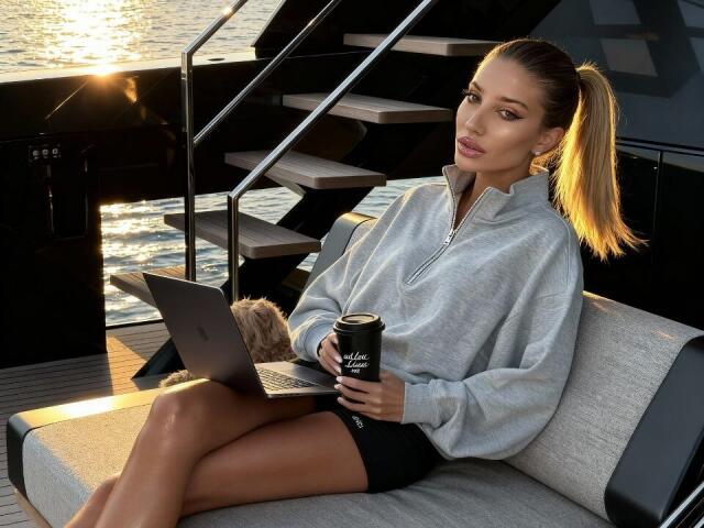 ElzaKomx zit op een grijze stoel, met gekruiste benen, en draagt een grijze hoodie en zwarte korte broek. Ze houdt een koffiekop en een laptop vast. zonlicht dat op het water in de achtergrond reflecteert, ontspannen pose.