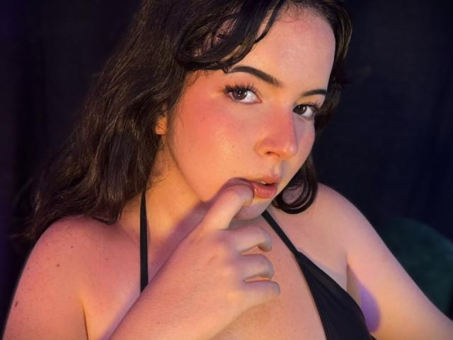 Close-up of AliinaFontaine, fair-skinned with dark wavy hair, biting her finger while looking seductively at the camera. She's wearing a black halter top, illuminated with pink and yellow light.