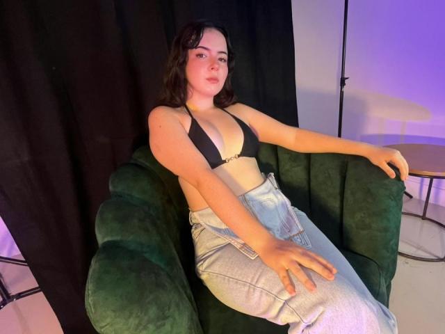 AliinaFontaine with dark hair, pale skin, and a curvy physique sits on a green armchair, wearing a black bra and shiny silver pants. Her hand rests on her crotch, exuding sexual tension.