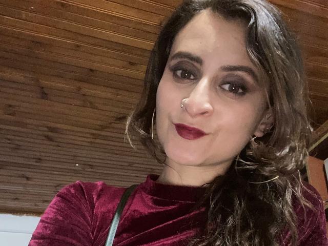 Nahaufnahme-Selfie von SaraVelvett, einer braunhaarigen Frau mit dunklem Make-up, rotem Lippenstift, einem Nasenring und Creolen, die trägt ein dunkelroter Rollkragenpullover. Hintergrund mit Holzboden und Decke. Leices Lächeln, suggestiver Blick..