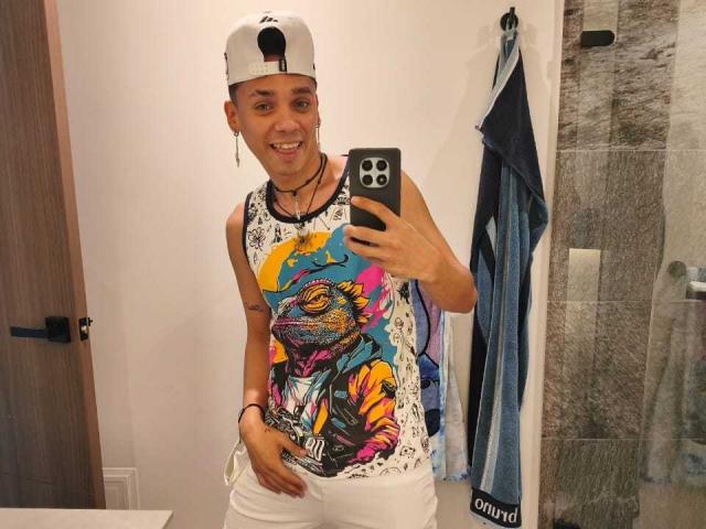 MaxxKing está em um banheiro, sorrindo para o espelho, segurando um telefone, vestindo uma camiseta com uma estampa colorida, calças brancas e boné usado de trás para frente. Uma toalha está pendurada à direita. Que fofa..