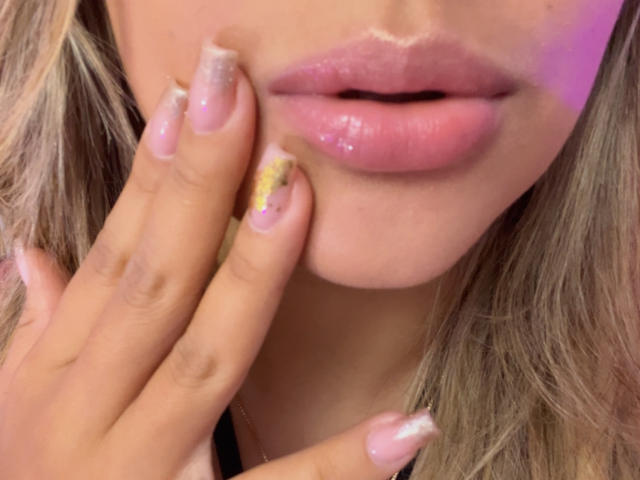 Primo piano del viso di SashaPolet, con particolare attenzione alle sue labbra rosa lucide e alla mano con le unghie scintillanti che la sfiora delicatamente sul mento. labbra.
i suoi capelli biondi incorniciano il suo viso, aggiungendo un'aria seducente..