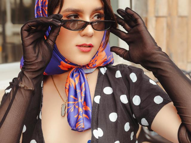 Göttin mit dunkelbrauner Haut, die ein schwarzes Top mit weißen Punkten, einen blauen Schal mit orangefarbenen Mustern, schwarze Spitzenhandschuhe trägt und dunkle Sonnenbrille. Hände, die eine Brille justieren, leichtes Lächeln, Hintergrund einer Straße..