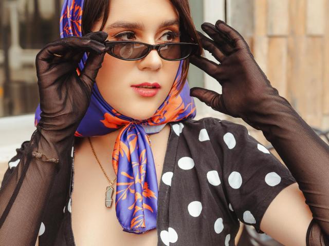GoddessBar con una camisa de lunares negros, una bufanda azul y naranja, guantes de malla negra, ajustándose gafas de sol negras, vistiendo de rojo pintalabios, collar colgante, piel clara, fondo urbano, ambiente sexy y seguro, primer plano detallado..