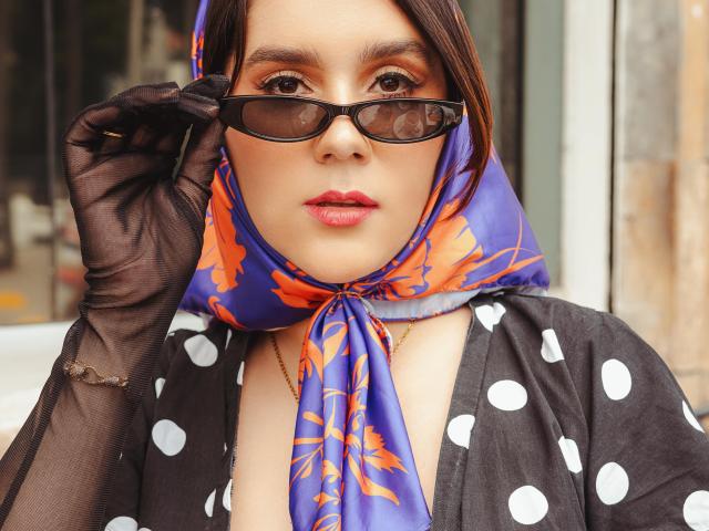 Close-up van GoddessBar, die een blauwe hoofddoek draagt met oranje bloemmotieven, een zwart gebreide vest met stippen en een zwarte kanten top. handschoenen, en past donkere zonnebrillen aan. Ze heeft rode lippenstift en een serieuze uitdrukking..
