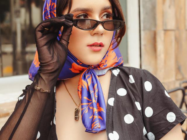 GoddessBar trägt einen blauen und orangefarbenen Blumen-Kopfschal, ein schwarzes Top mit Punkten und transparente schwarze Handschuhe und justiert etwas. Sonnenbrillen. Sie trägt eine Halskette mit einem kleinen Anhänger. Verdammt sexy..