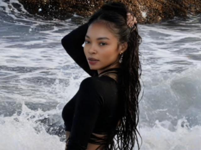 Fotografia de SwettBrown, uma mulher asiática atraente com longos cabelos pretos e molhados, em pé no oceano. Ela está olhando por cima do ombro, com a mão no cabelo, enquanto as ondas quebram atrás dela. Muito atraente..