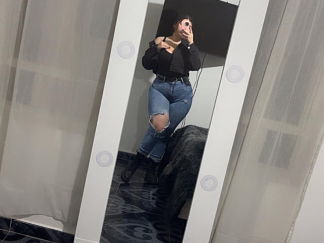 VenusxMusa se toma un selfie frente a un espejo de cuerpo entero con luces brillantes. Ella lleva una sudadera negra con capucha, jeans azules rotos y botas negras, y está señalando a la cámara. Pantalones vaqueros ajustados y muy atractivos..