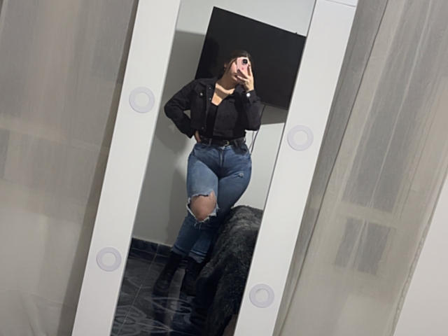 VenusxMusa, een aantrekkelijke vrouw met donker haar, een zwarte top en gescheurde blauwe jeans, staat voor een verlichte spiegel, met haar hand gezicht bedekt, zwarte bontjas op de vloer, intense spiegeling..