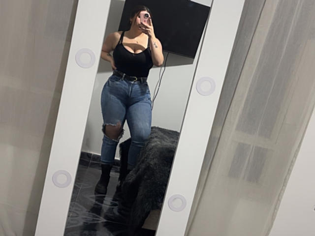 VenusxMusa, a curvy woman with large breasts, stands in front of a mirror, holding a phone, wearing a black tank top and ripped jeans, showing off her hourglass figure. Reflection highlights her sexy pose.