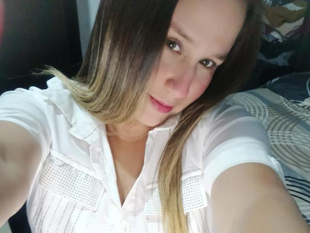 Autoritratto ravvicinato di AlexaHotSeins, una donna con lunghi capelli castani, che indossa una camicetta bianca e guarda direttamente la fotocamera. con un'espressione leggermente seducente, luce naturale intensa..