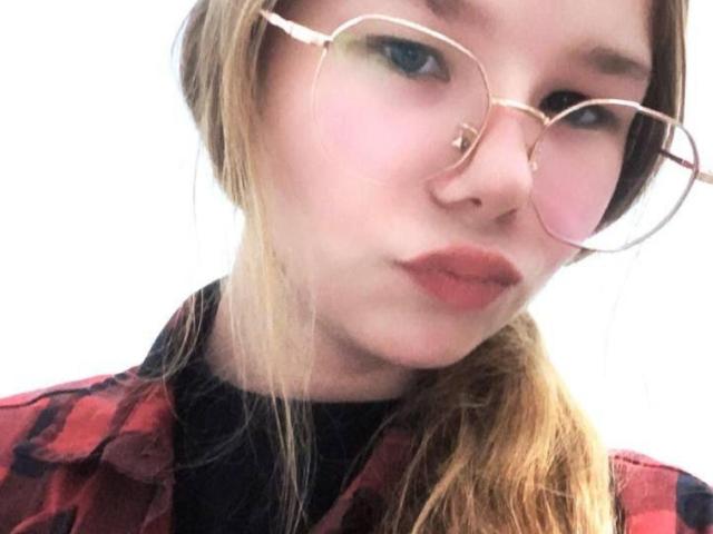 Nahaufnahme von JohanaDelduca mit heller Haut, blauen Augen und roten Lippen. Sie trägt eine runde Brille und ein rotes Karohemd über einem schwarzen Rollkragenpullover. Gerade Pony, hellbraunes Haar. Verdammt sexy..