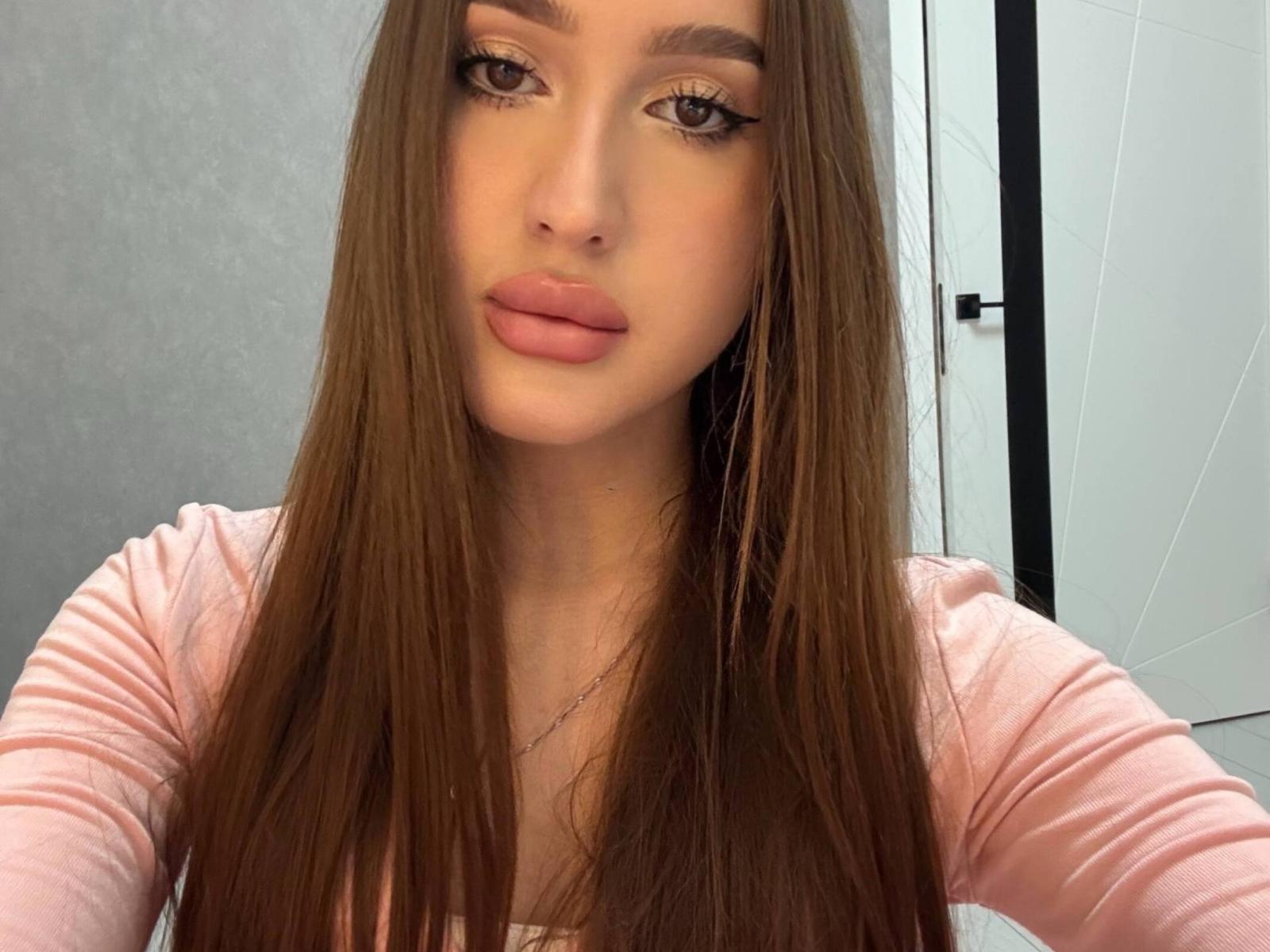 Primo piano di SensuallMaya, una giovane donna con lunghi capelli castani, labbra piene e un trucco pesante per gli occhi, che indossa un abito rosa. Maglia a maniche lunghe, che guarda direttamente l'obiettivo con un'espressione leggermente provocante..