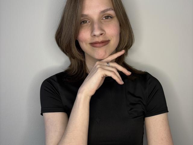 Foto van Lourie Hegner met een lichte huid en bruin haar, die een zwarte blouse draagt en poseert met één hand in de buurt van haar kin. lichte glimlach, lichte blos, zachte belichting, informeel en enigszins flirterig.