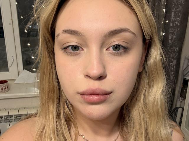 Photo en gros plan de RubiCharm, une jeune femme à la peau claire, aux cheveux bruns clairs et à l'expression neutre. Elle a de petits sourcils, un trait d'eye-liner discret, et des lèvres d'un rose clair. Guirlandes lumineuses en arrière-plan.
