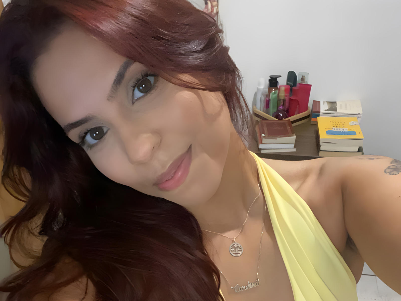 Fotografía de primer plano de HsiuGartman, una mujer con cabello rojo largo y ondulado, que lleva un top amarillo con tirantes, sonriendo ligeramente, de color azul. ojos, maquillaje ligero, collar, tatuaje en el hombro izquierdo, fondo desordenado. Muy atractivo..