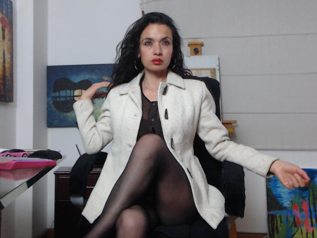 AfroditaRgenis, seated in a black chair, wearing a white coat over a black dress, black stockings, with legs crossed, hand near face, exuding confident, sexy vibe, slight smirk, modern art background.