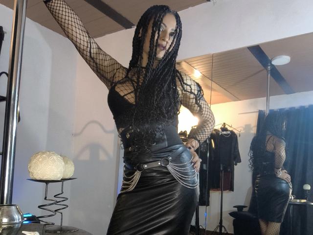 DivaMarie, standing in a dimly lit room, wearing a black fishnet top and leather skirt, arms raised, showing off long braids and confident pose, with a sexy, provocative vibe.
