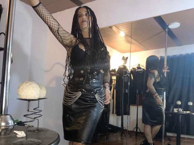 DivaMarie with long black dreads, wearing a black fishnet top and leather skirt, stands confidently with one arm raised. Reflection in mirror shows her sexy backside. Dimly lit room with lamps.