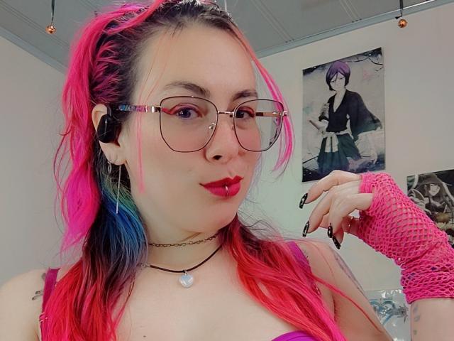 Foto ravvicinata di NatashaLau con capelli rosa e blu, che indossa occhiali, rossetto rosso, canottiera nera, guanto a rete rosa. Collana choker. Lo sfondo include poster di anime. Una posa incredibilmente sexy e sicura di sé..
