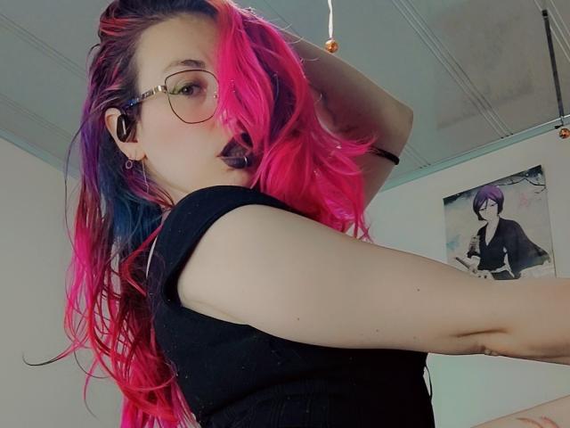 NatashaLau with pink and black hair, wearing glasses and a black top, looking over her shoulder, hand in hair. Background: white wall, poster of another person. Slight sexual tension.