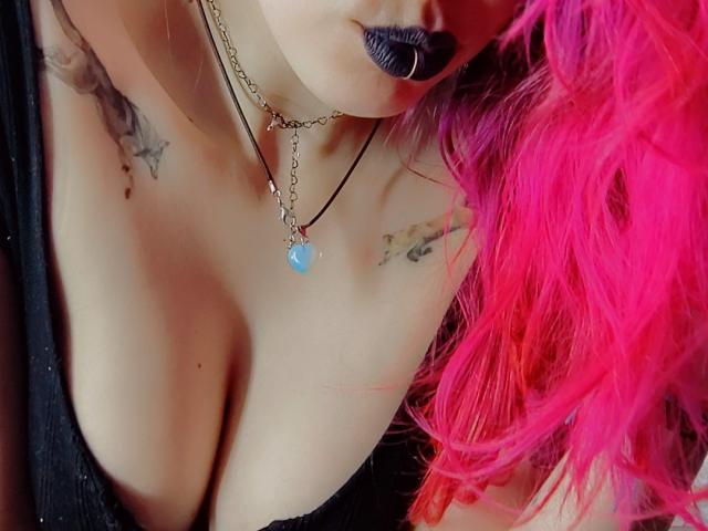Gros plan de la poitrine et du cou de NatashaLau, montrant le décolleté, du rouge à lèvres noir, des cheveux rose vif, des tatouages sur les clavicules. Débardeur noir, collier avec pendentif bleu. La tension sexuelle dégagée par son rouge à lèvres noir et sa poitrine dénudée..