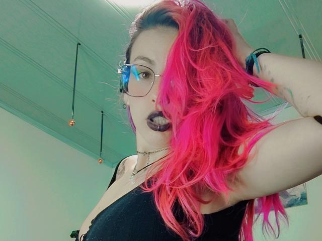 NatashaLau, avec ses cheveux rose vif, portant du rouge à lèvres noir et des lunettes, pose, une main dans ses cheveux, en haut noir apparent. Décolleté, peau claire, intérieur, plafond moderne avec des luminaires suspendus. Incroyablement sexy..