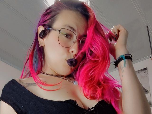 Nahaufnahme von NatashaLau mit leuchtend rosa und schwarzen Haaren, die schwarzen Lippenstift, eine Brille und ein schwarzes Oberteil trägt, das einen Schulter frei lässt. oben. Sie zupft an ihren Haaren, hat ein Tattoo am Handgelenk und trägt eine schwarze Halskette. Verdammt heiß.
