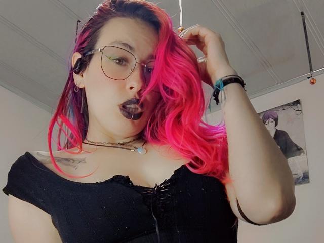 NatashaLau, con i capelli rosa e neri, indossa occhiali e una maglietta nera con una spalla scoperta, ha una mano tra i capelli, con rossetto nero. tatuaggi sul petto, collana bianca, sguardo intenso, tensione sessuale, stile selfie.