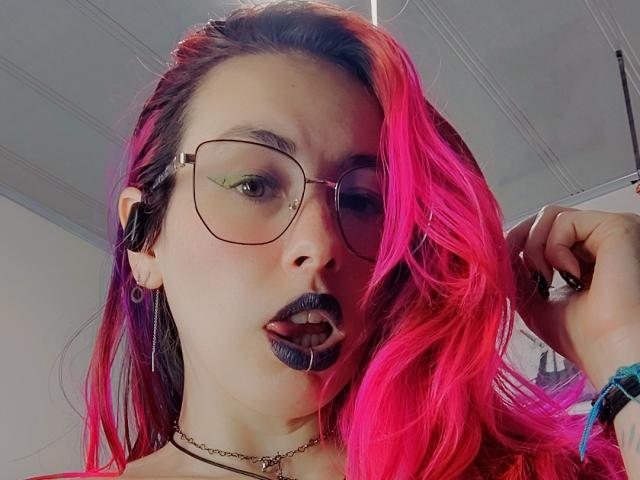 Gros plan de NatashaLau avec des cheveux roses éclatants, du rouge à lèvres noir et des lunettes. Elle se tient les cheveux, regardant l'appareil photo avec une bouche légèrement ouverte. Portant un collier ras du cou noir et un bracelet..