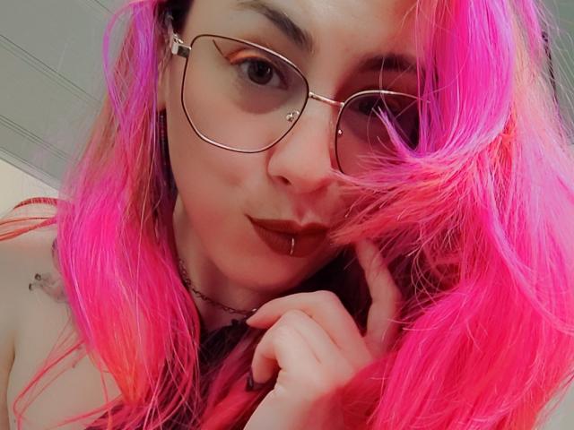 Nahaufnahme-Selfie von NatashaLau mit leuchtend rosa Haaren, großen, runden Brillen und einem verführerischen Ausdruck. Sie berührt ihr Kinn mit einer Hand, ihre Lippen sind leicht geöffnet, und sie strahlt eine sexuelle Spannung aus..