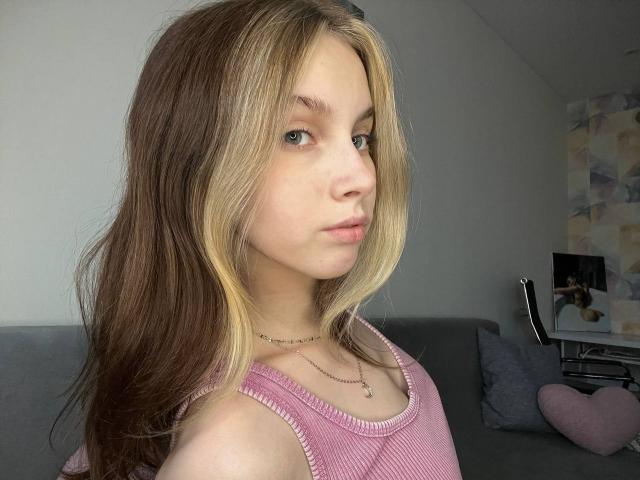 Fotografia de Ione Trosper com cabelos castanhos longos e mechas loiras, vestindo uma camiseta rosa, olhando por cima do ombro. com uma expressão ligeiramente séria. Luz natural suave, ambiente interno..