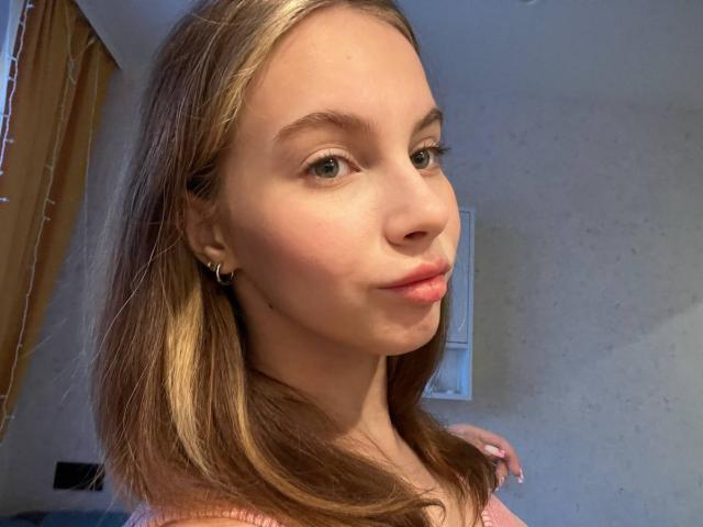 Close-up foto van IoneTrosper met lichtbruin haar, lichte huid en subtiele make-up. Ze heeft een lichte grijns en draagt een roze top. Zacht licht accentueert haar jukbeenderen, waardoor ze een sexy en ontspannen uitstraling heeft..