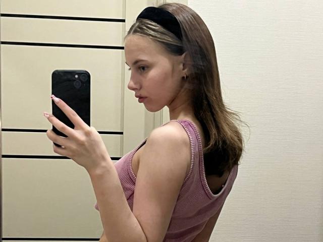 Ione Trosper, met bruin haar dat tot op haar schouders reikt, draagt een roze top van gaas en houdt een zwarte telefoon vast, terwijl ze een selfie maakt. Ze heeft een geconcentreerde uitdrukking, en haar slanke, atletische lichaam is duidelijk zichtbaar..