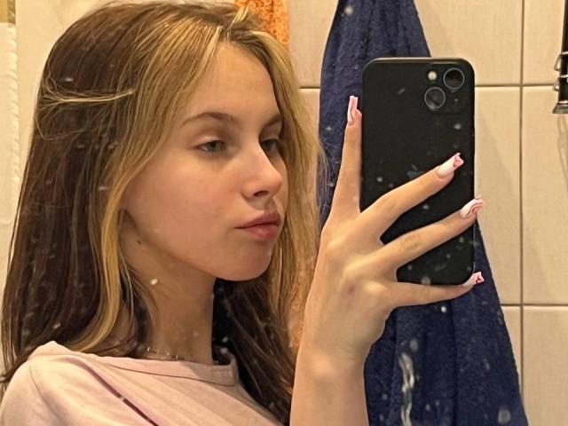 Ione Trosper, met nat bruin haar, volle lippen en roze nagels, houdt een zwarte telefoon vast en maakt een selfie in een betegelde badkamer. Ze draagt een lichtroze shirt, op de achtergrond is een blauwe handdoek te zien..