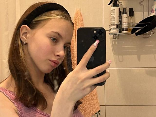 Ione Trosper, con cabello castaño y una diadema negra, vestida con una camiseta rosa, se toma un selfie en un baño. Ella sostiene un teléfono negro, con botellas de champú en un estante detrás de ella..