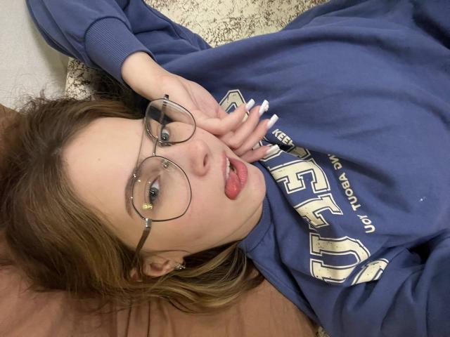 IoneTrosper lying on her back, wearing a blue hoodie with "USC" text, glasses, and red lipstick. Her hand touches her face, brown hair, carpet beneath her, casual sexy pose.