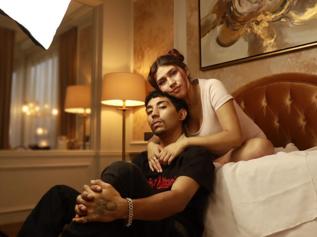 Fotografía de Anyelin y Mark sentados en una cama, con la cabeza de ella apoyada en el hombro de él, ambos mirando a la cámara. Iluminación cálida, cabecera de cuero marrón, sábana blanca. Ella lleva una camisa blanca, él lleva una camisa negra..