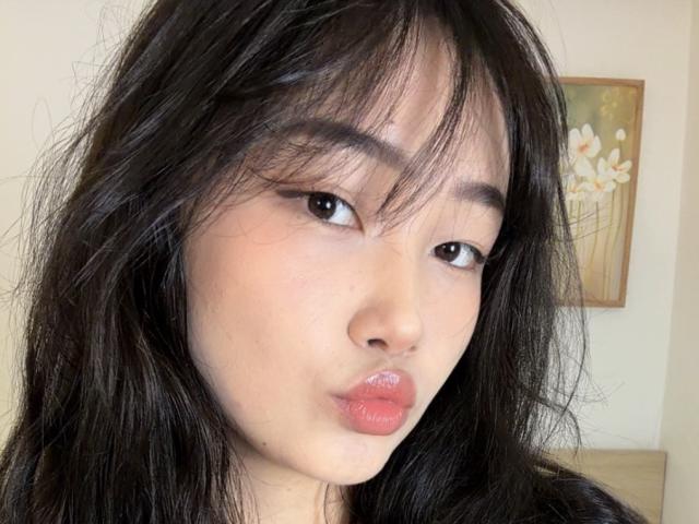 Close-up of PinkyBlooom with black, slightly messy hair, pouty lips, and Asian features. PinkyBlooom has a suggestive expression, slightly parted lips, and dark eyes. Framed flower painting in background.